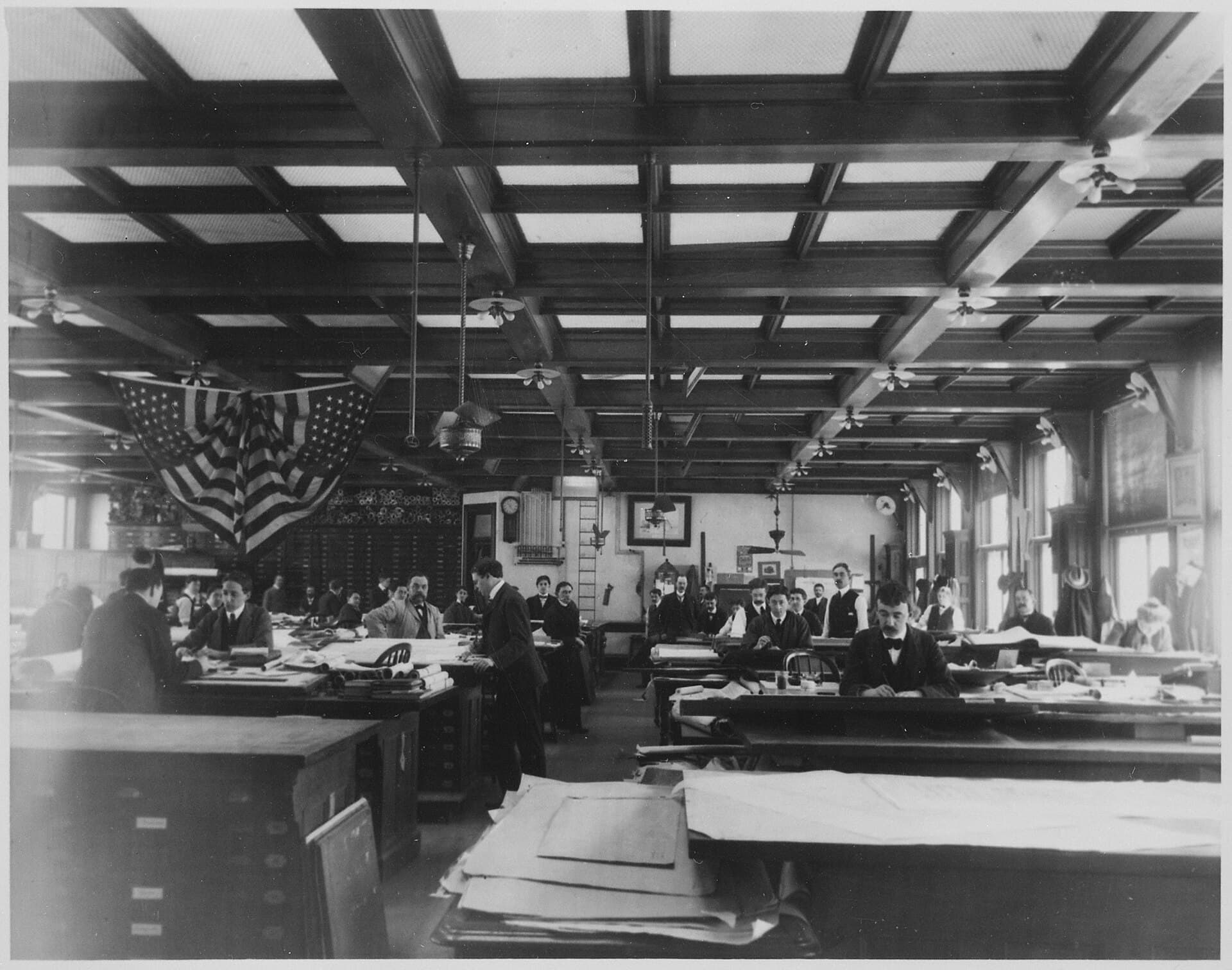 Architects and draftsmen working at rows of drafting tables covered in drawings, in a skylit drafting room